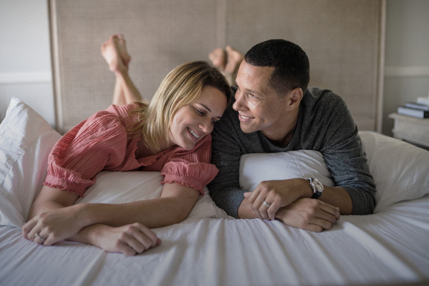 photo of a smiling couple on bed with the feet resting in the headboard. the girl wearing a diamond ring and resting his head on the guy's shoulder wearing a wedding ring and a watch