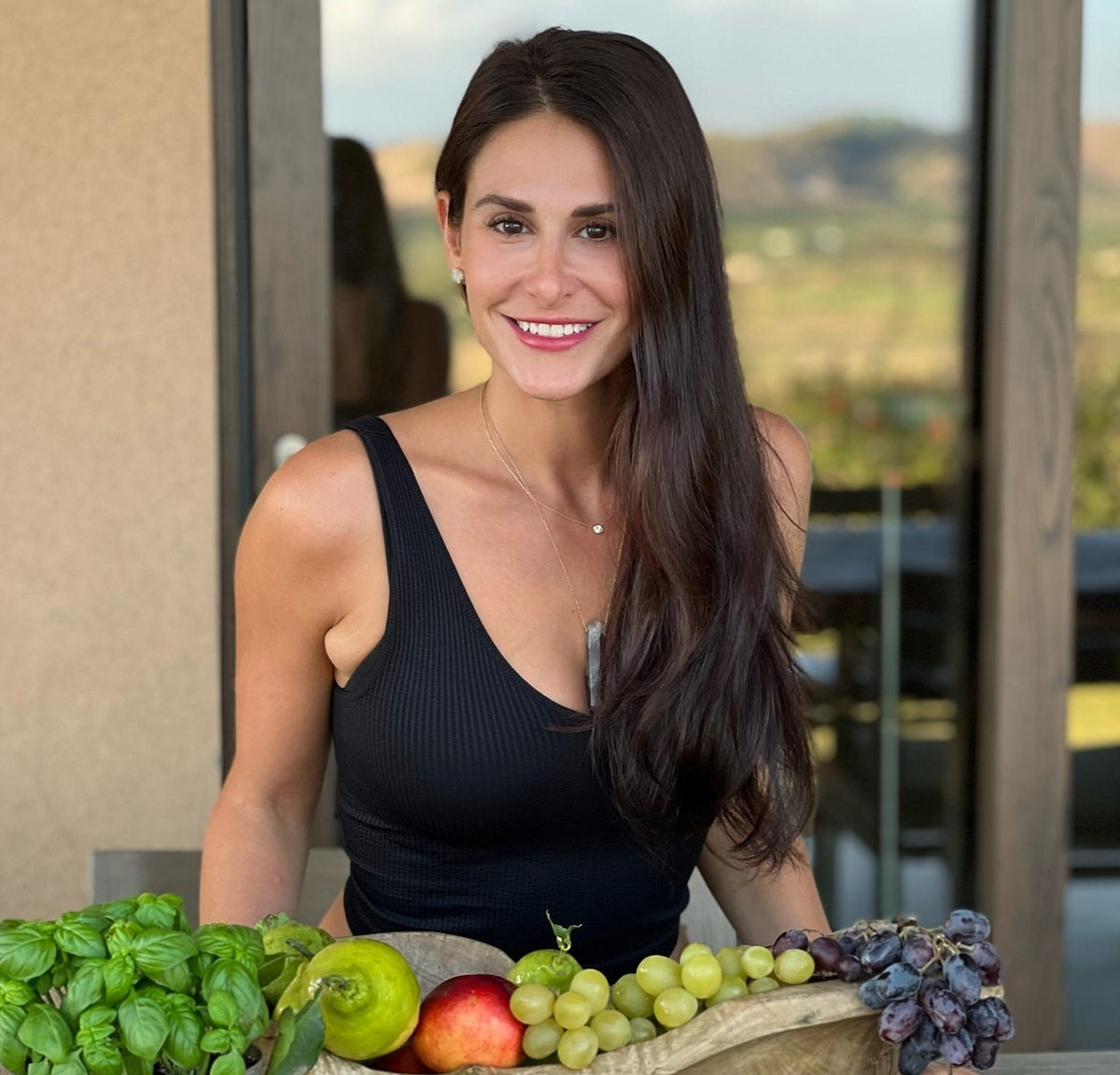 A woman in a black tank top smiles at the camera, with fruits like apples and grapes nearby.