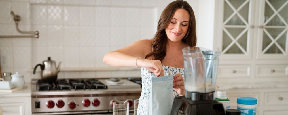 Smiling woman in a bright kitchen preparing a smoothie with a blender, surrounded by WeNatal supplements and other wellness products, emphasizing health, nutrition, and preconception wellness