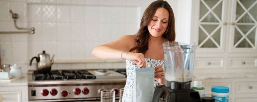 Smiling woman in a bright kitchen preparing a smoothie with a blender, surrounded by WeNatal supplements and other wellness products, emphasizing health, nutrition, and preconception wellness.