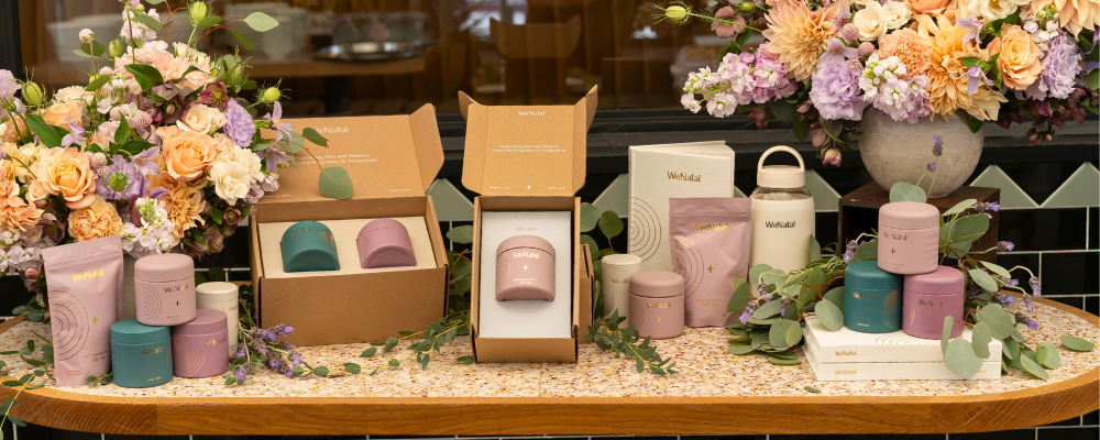 A display of WeNatal products including floral arrangements, pink containers with gold text, and a jar of cream, highlighting elegant packaging.