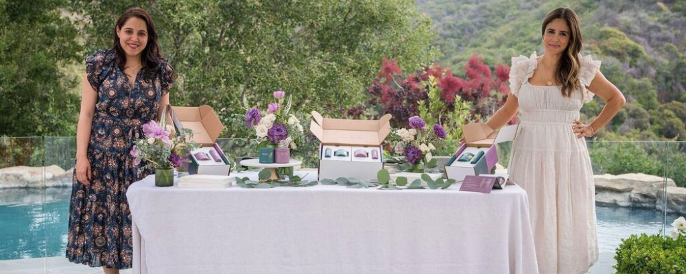 Ronit and Vida standing by a beautifully decorated table showcasing WeNatal prenatal supplements, surrounded by flowers and a serene outdoor setting, symbolizing partnership and wellness. - WeNatal