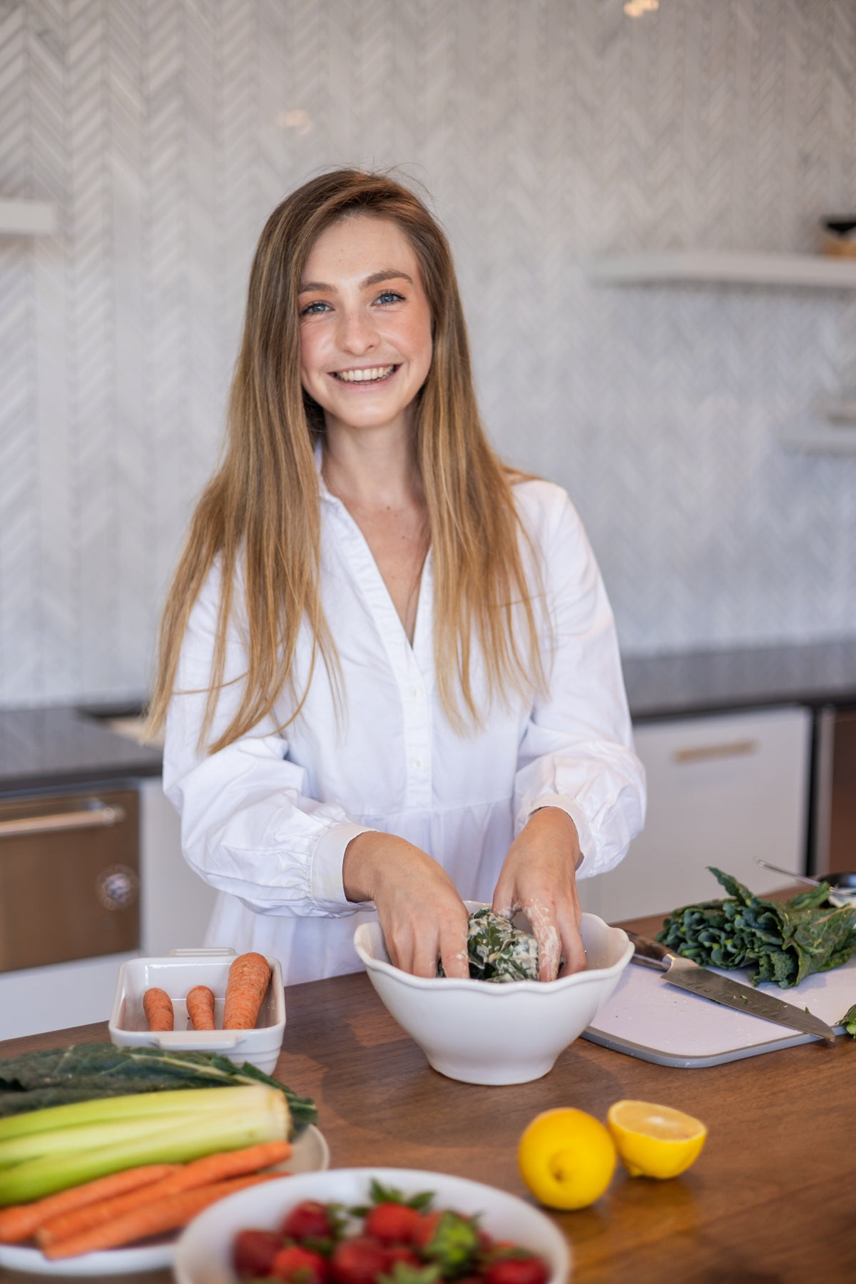 A woman smiling at the camera in a kitchen setting with fresh vegetables, including carrots and lemons, displayed nearby.