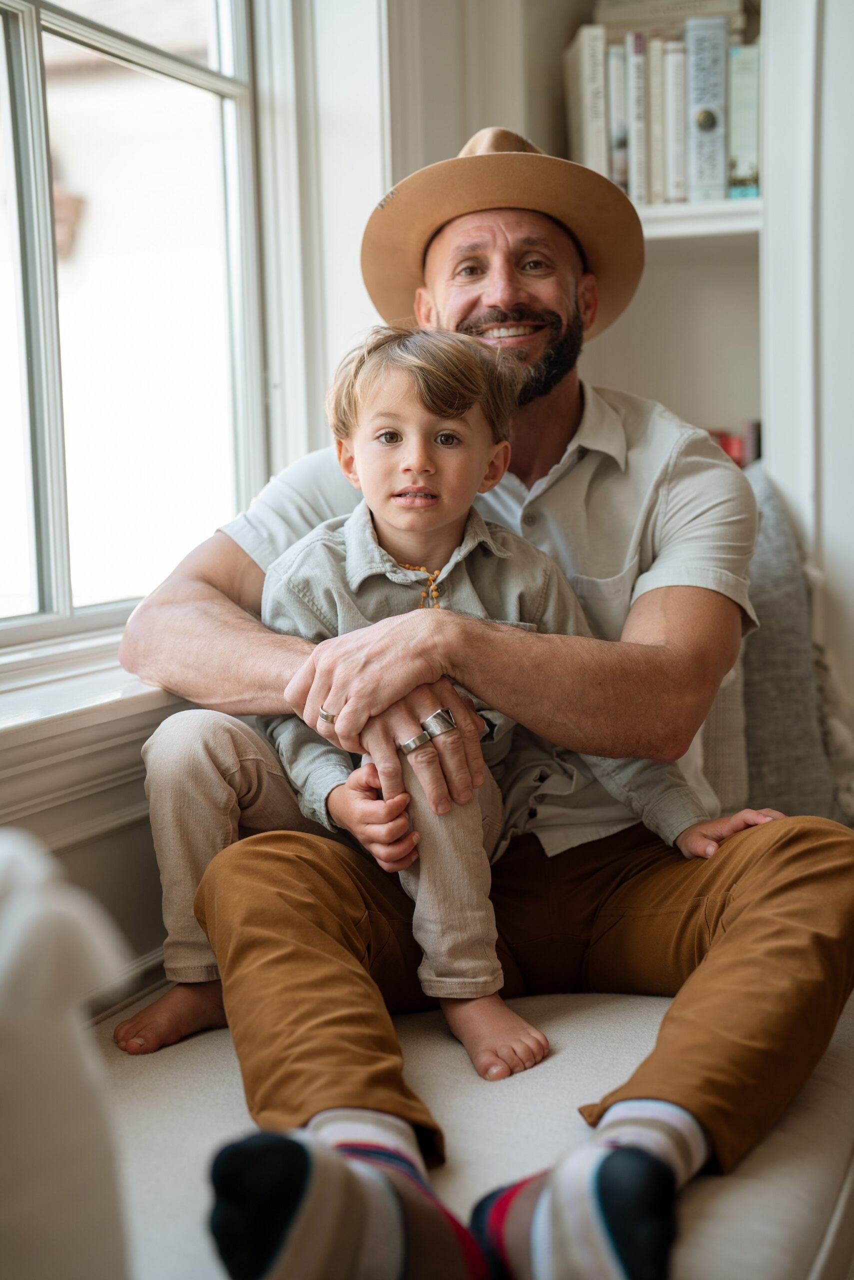A man wearing a hat sits on the floor, holding a smiling toddler, capturing a moment of comfort and connection indoors