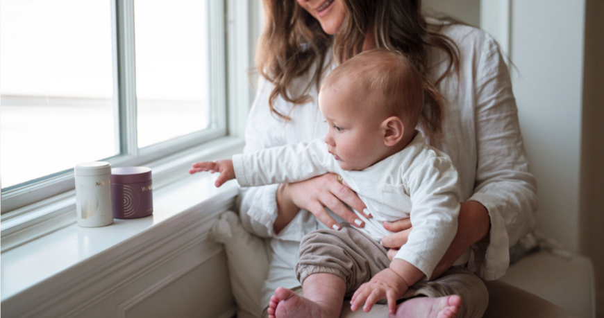 Mother holding her baby on her lap as the infant reaches for WeNatal for Her and Omega DHA+ supplement containers, highlighting essential prenatal nutrients for pregnancy and postpartum health.