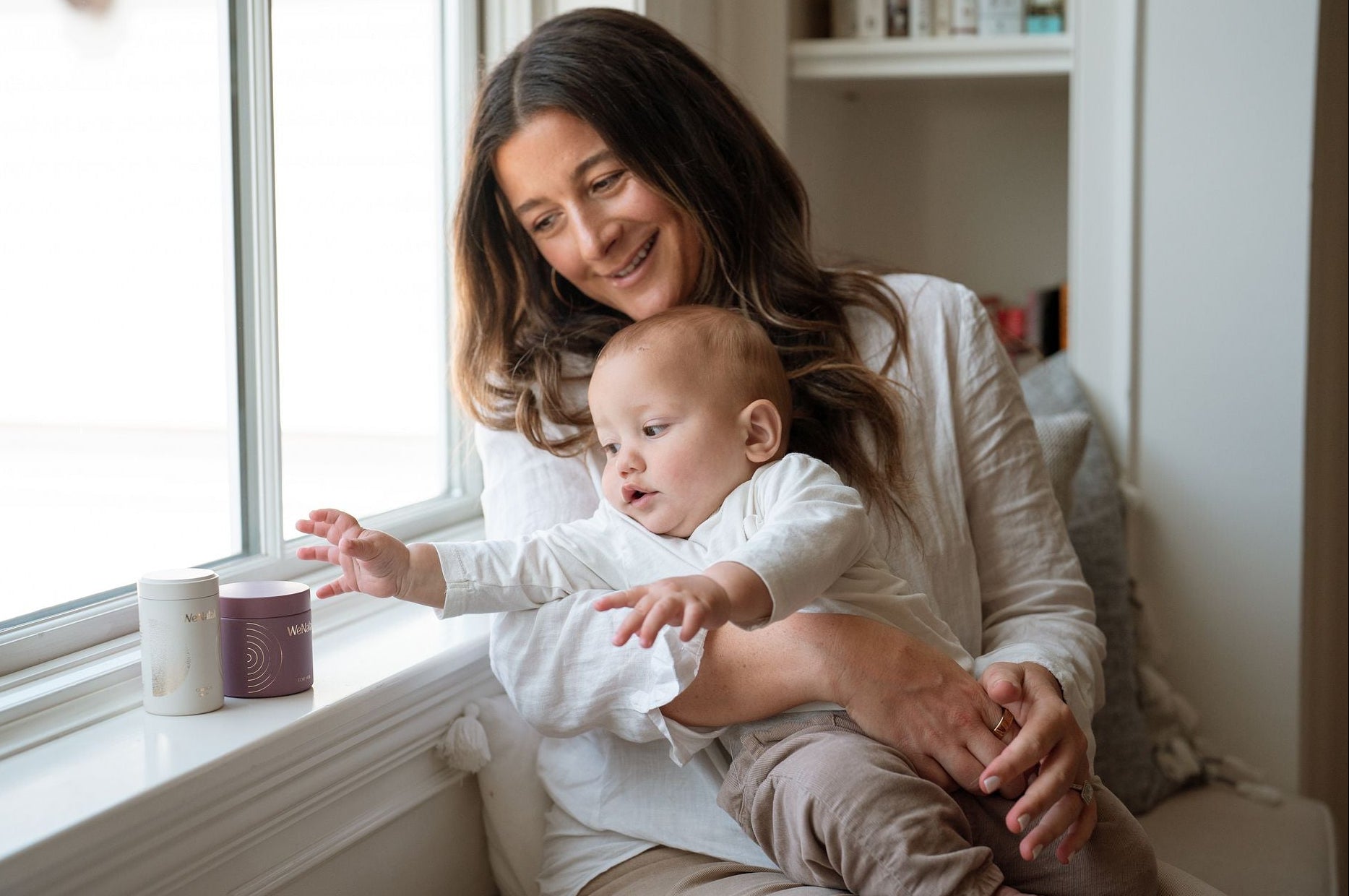 woman holding baby on her lap near sunny window with baby reaching for supplement jars - WeNatal prenatal