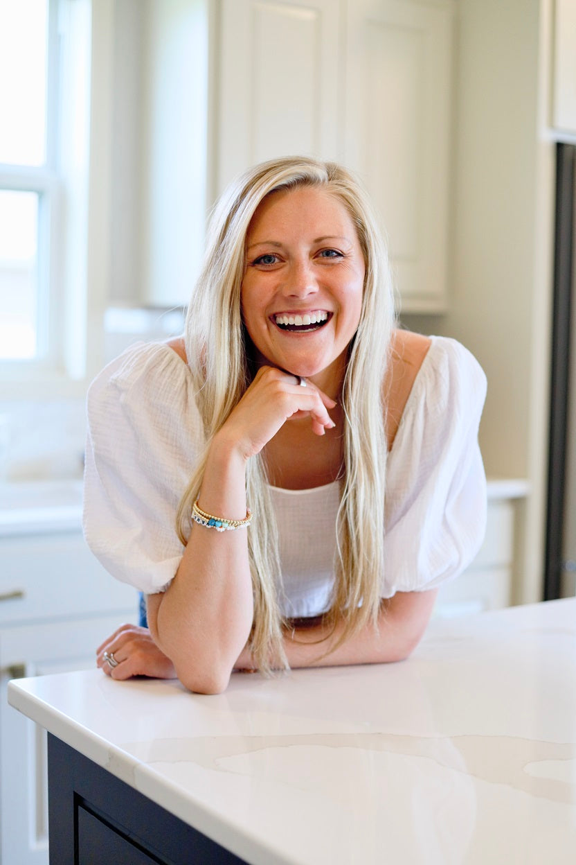 A woman smiling indoors, her hand on a white counter, wearing a bracelet, reflecting a positive and nurturing lifestyle associated with prenatal care.