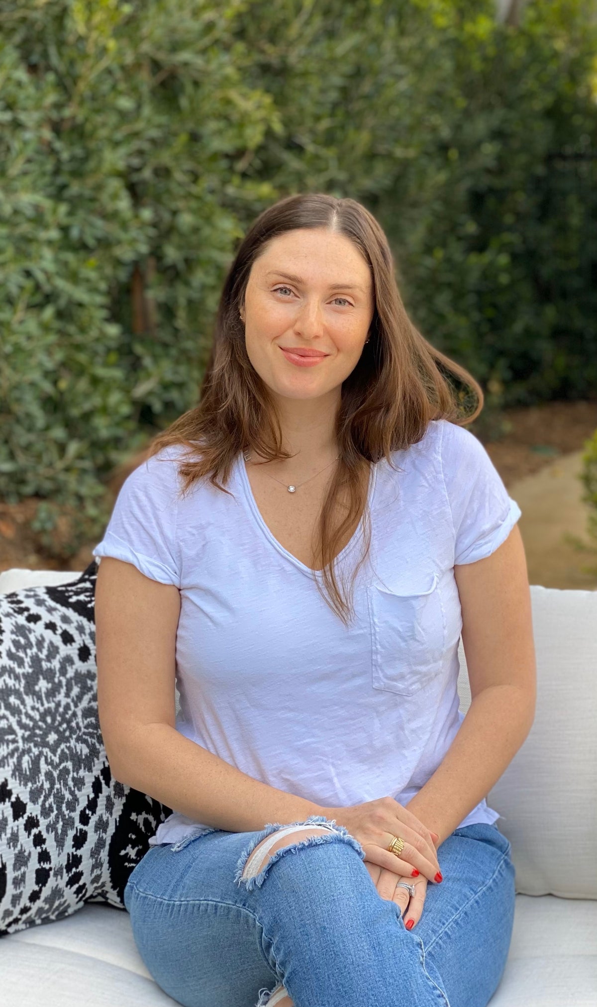 A woman in jeans with a torn knee sits on a bench outdoors, smiling at the camera.