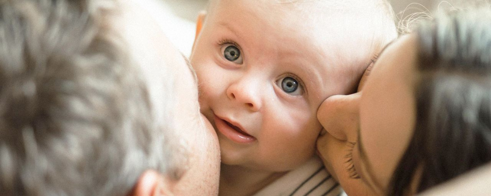 A mother and a father kissing the cheek of a young  baby with blonde hair and blue eyes - WeNatal