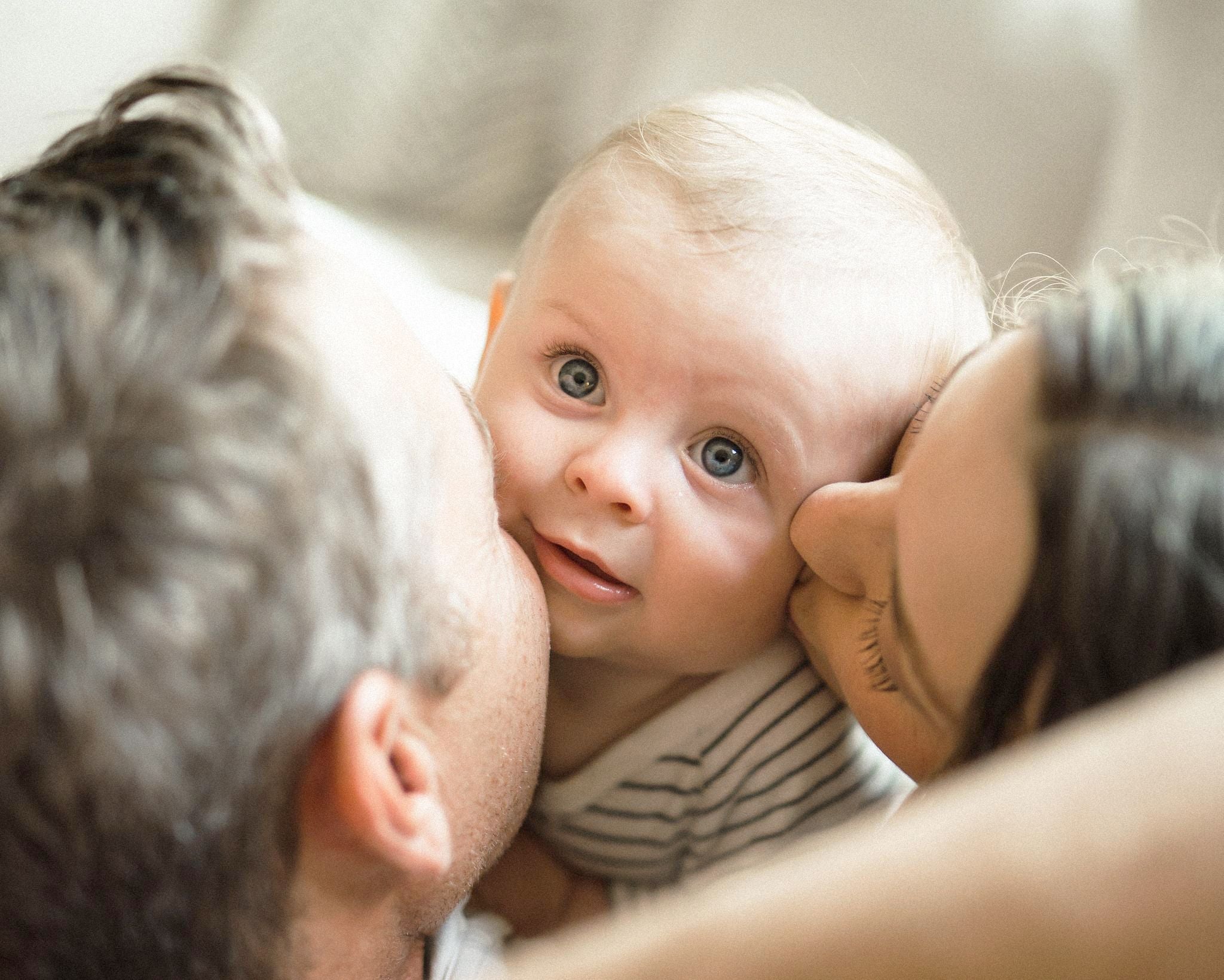 A mother and a father kissing the cheek of a young  baby with blonde hair and blue eyes - WeNatal
