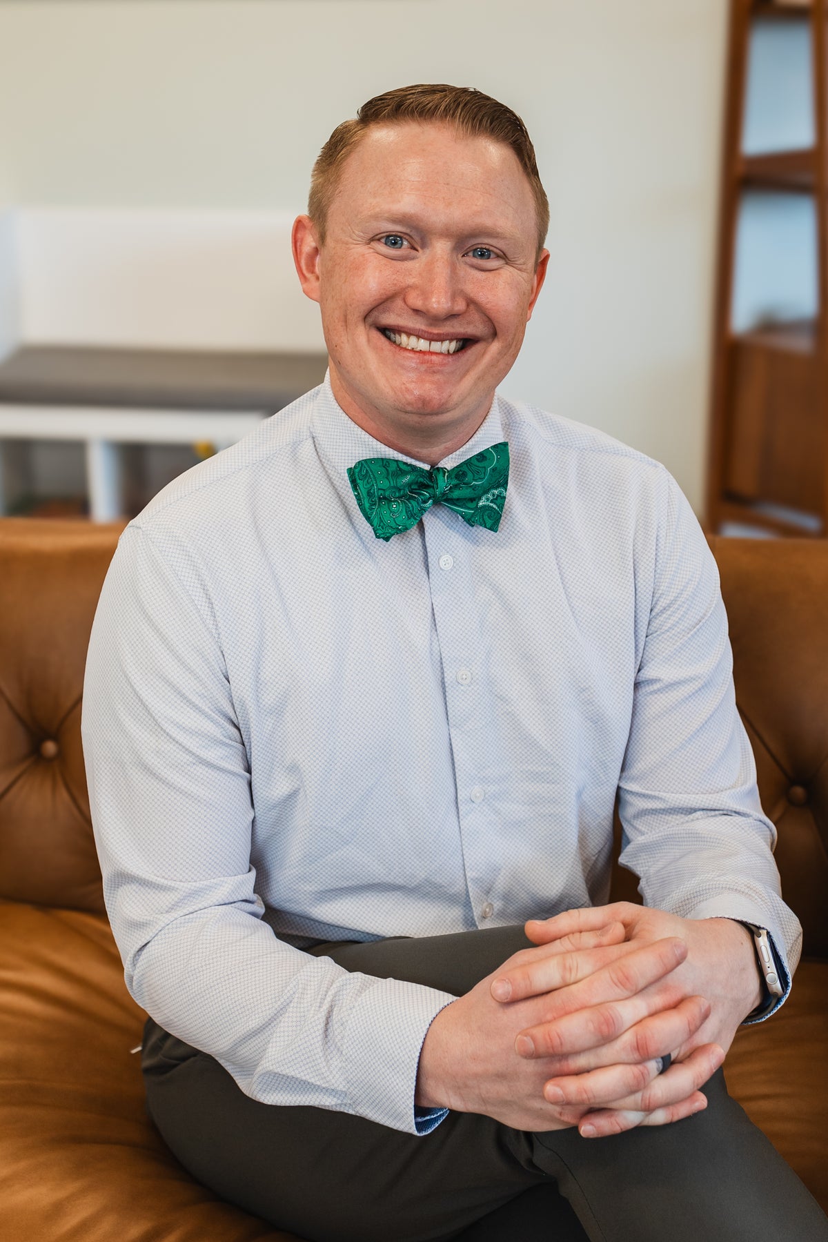 Man wearing a dress shirt and green bow tie with white and blue dots, smiling at the camera indoors.