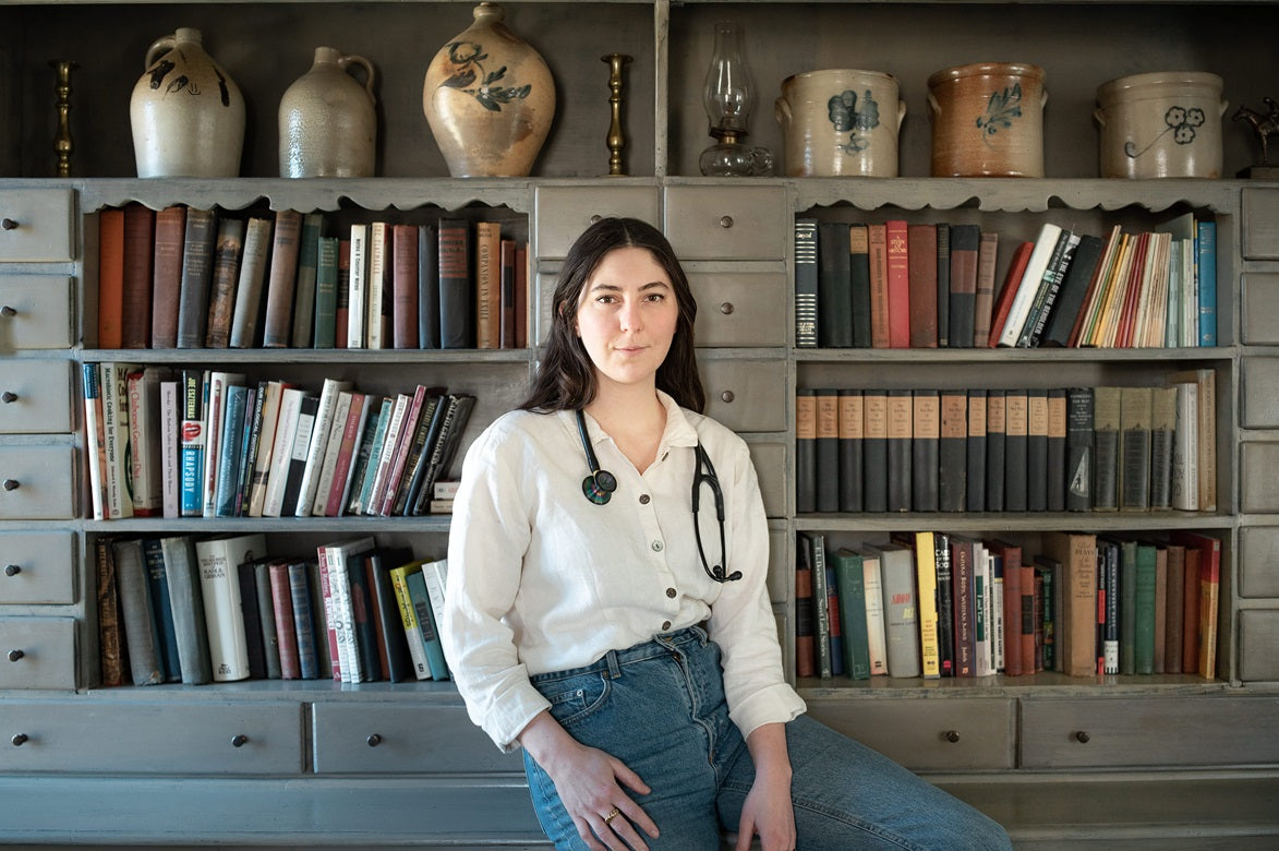 A woman sits in front of a bookshelf, surrounded by various vases and jugs, reflecting a serene indoor setting.