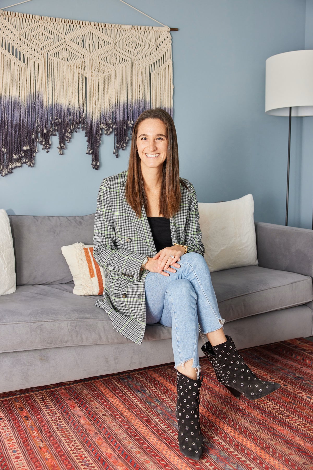 A woman smiling while sitting on a couch, wearing black boots, with an orange-striped pillow and a lamp visible in the background.