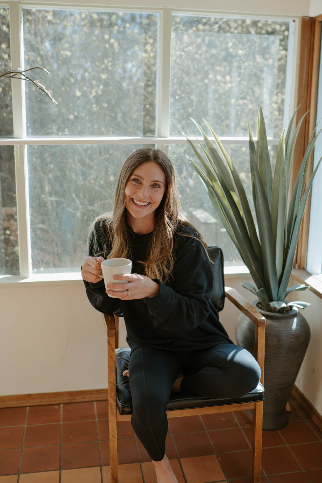 A woman sits in a chair holding a cup, smiling, next to a potted plant, indoors.