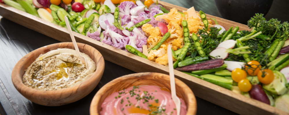 Colorful vegan platter with fresh vegetables including purple cauliflower, golden cherry tomatoes, asparagus, radishes, and broccolini served with hummus and beetroot dip, supporting plant-based nutrition for reproductive health - WeNatal prenatal