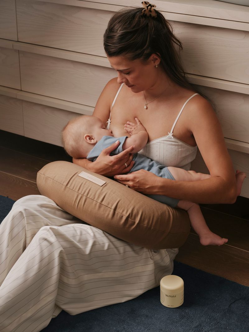 Mother breastfeeding her baby on a nursing pillow while sitting on the floor, with a jar of WeNatal Milk + Mood nearby - WeNatal Milk + Mood