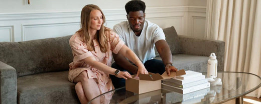 A man and woman sit on a couch, smiling while opening a box, possibly containing prenatal supplements from WeNatal.