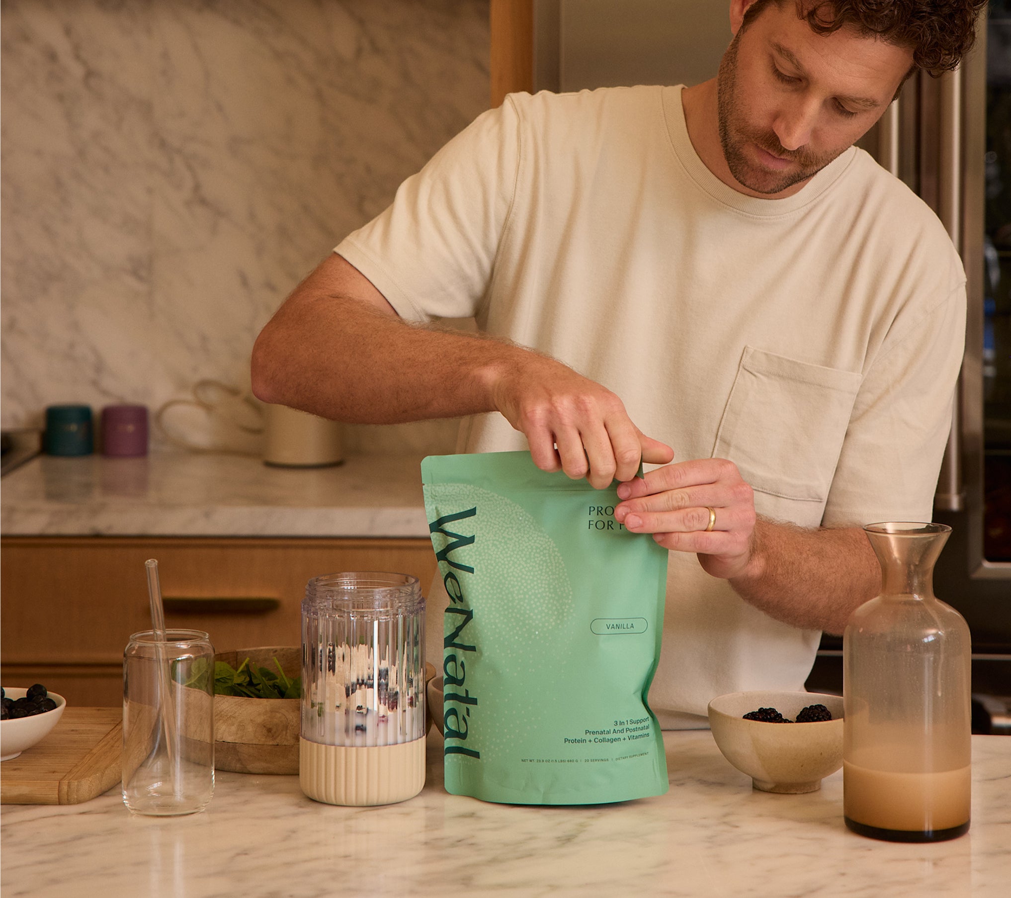 Person preparing a smoothie with WeNatal Prenatal Protein+ for Him, a 3-in-1 supplement pouch, featuring prenatal nutrition and nutrient support, alongside berries and leafy greens on a kitchen countertop