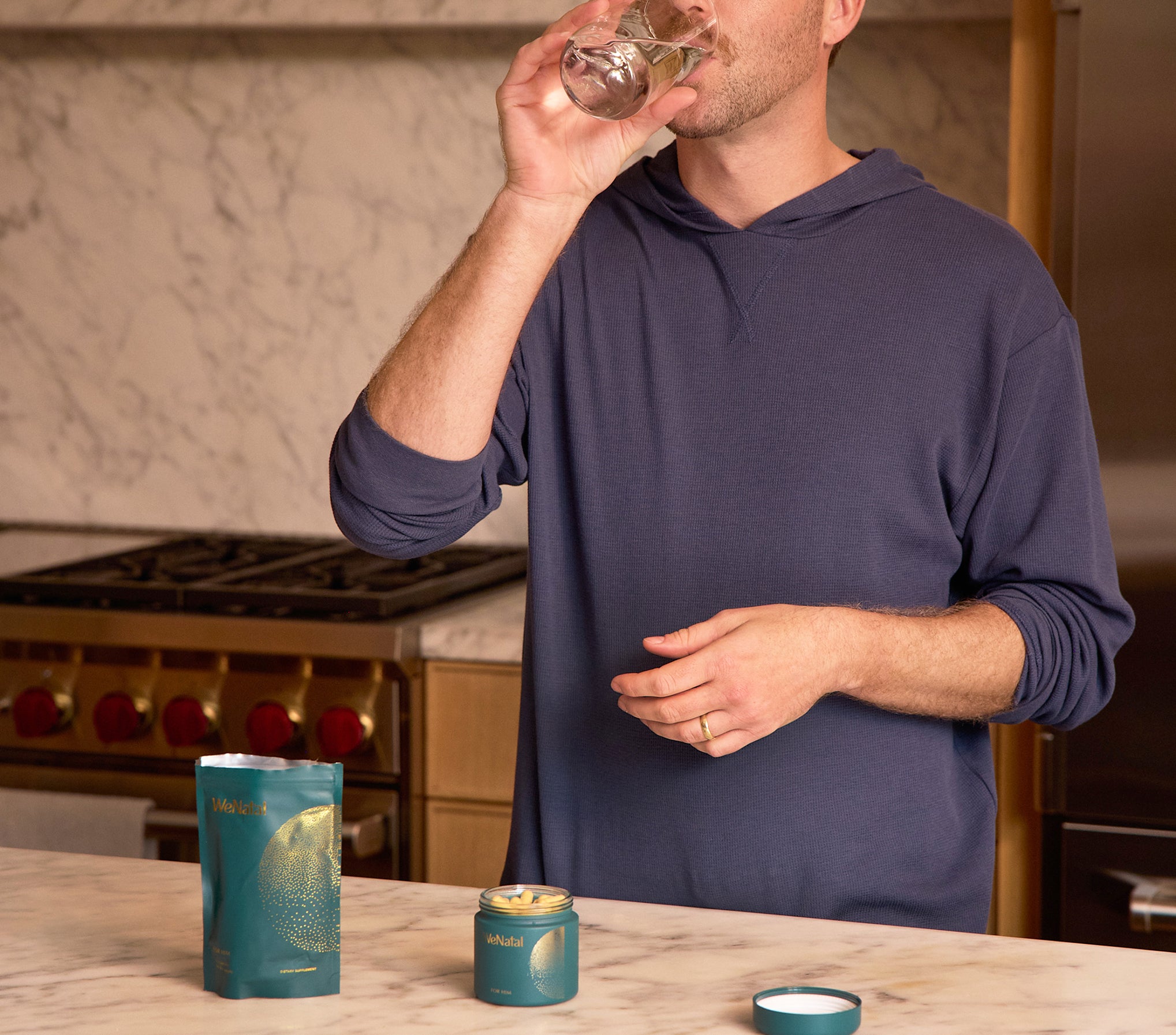 Man drinks beside WeNatal For Him Welcome Kit, showcasing a refill pouch and open jar with capsules, offering daily prenatal nutrient and fertility support