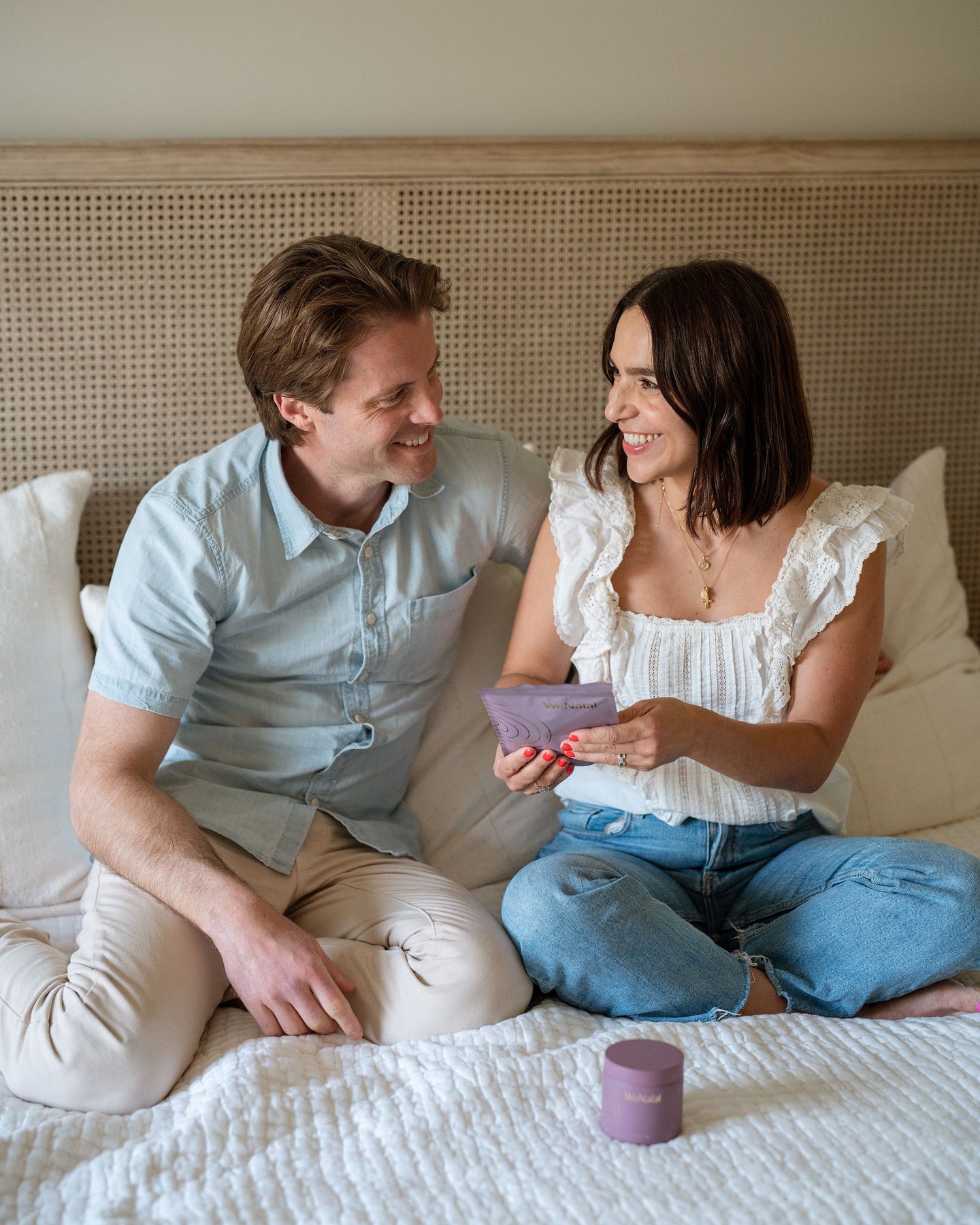 photo of a couple smiling at each other while sitting on their bed while the girl is carrying a refill pouch of WeNatal prenatal supplement for Her and the For Her glass jar placed on top of the bed too near the girl