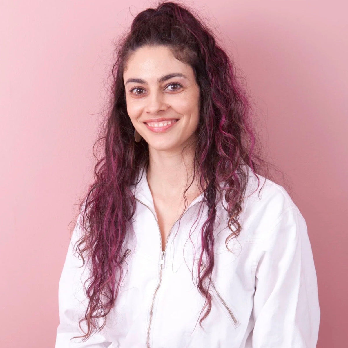 A smiling woman with long, wavy magenta-pink hair wearing a white zip-up jumpsuit, standing against a solid pink background.

