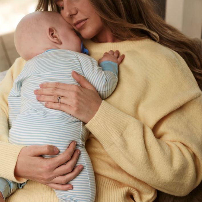A woman in a pale yellow ribbed sweater holds a baby wearing a light blue and white striped one-piece with solid blue cuffs.