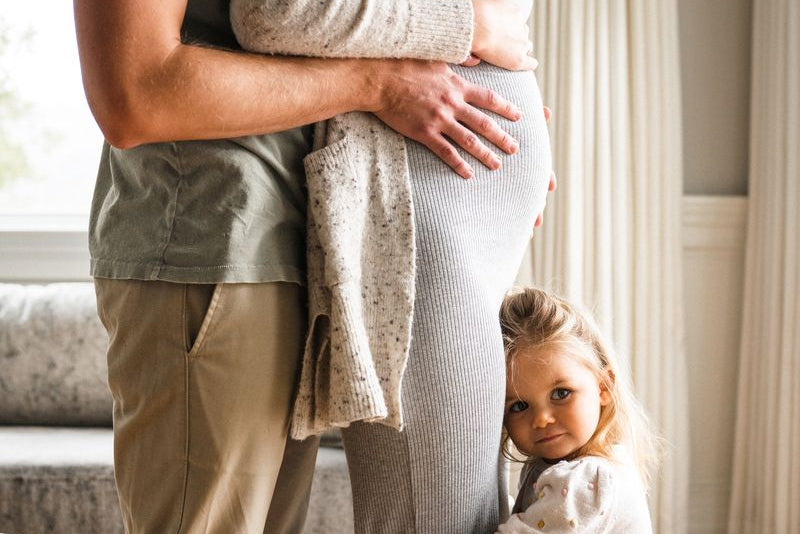 husband standing beyond wife both holding her pregnant stomach with toddler hugging mom’s legs - WeNatal Prenatal - WeNatal male fertility
