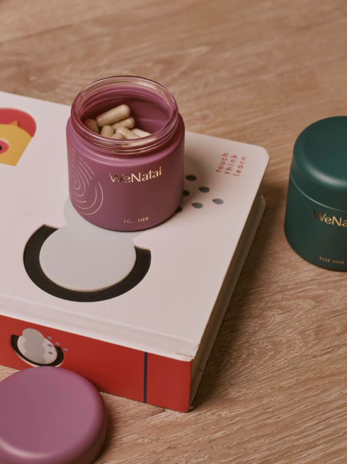 An open WeNatal For Her jar of capsules on a children's toy box, with a WeNatal For Him jar beside it