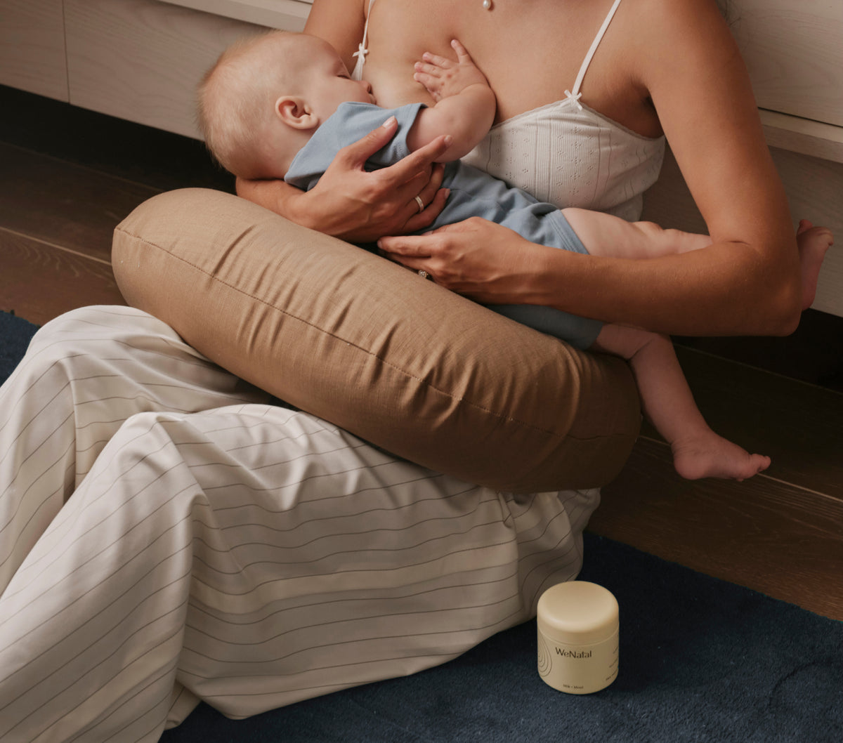 A mother breastfeeding her baby while sitting on the floor with a nursing pillow, a WeNatal Milk + Mood jar beside her