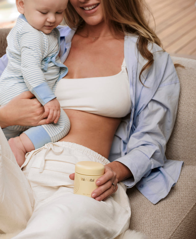 A smiling mother holding her baby and a WeNatal Milk + Mood jar while sitting on a couch