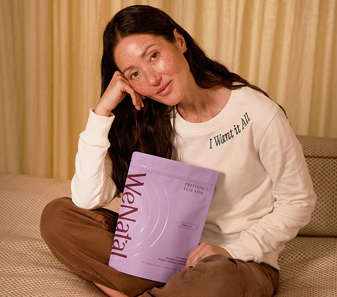 Woman in WeNatal Sweatshirt - 2026 holds a WeNatal Protein for Her pouch, illustrating daily prenatal nutrition and fertility support within a warm, cozy setting