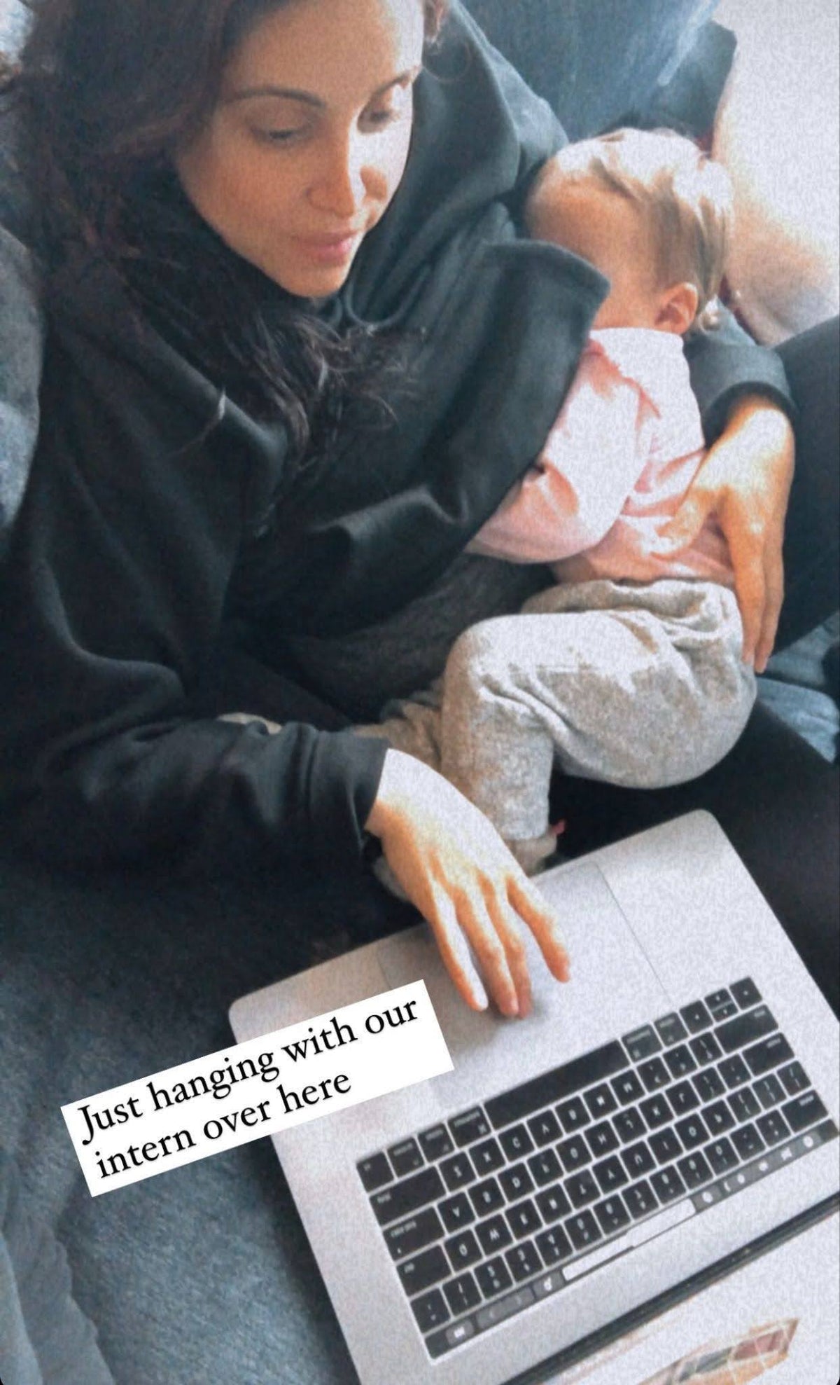 A woman working on a laptop while holding a toddler, captioned 'Just hanging with our intern over here'