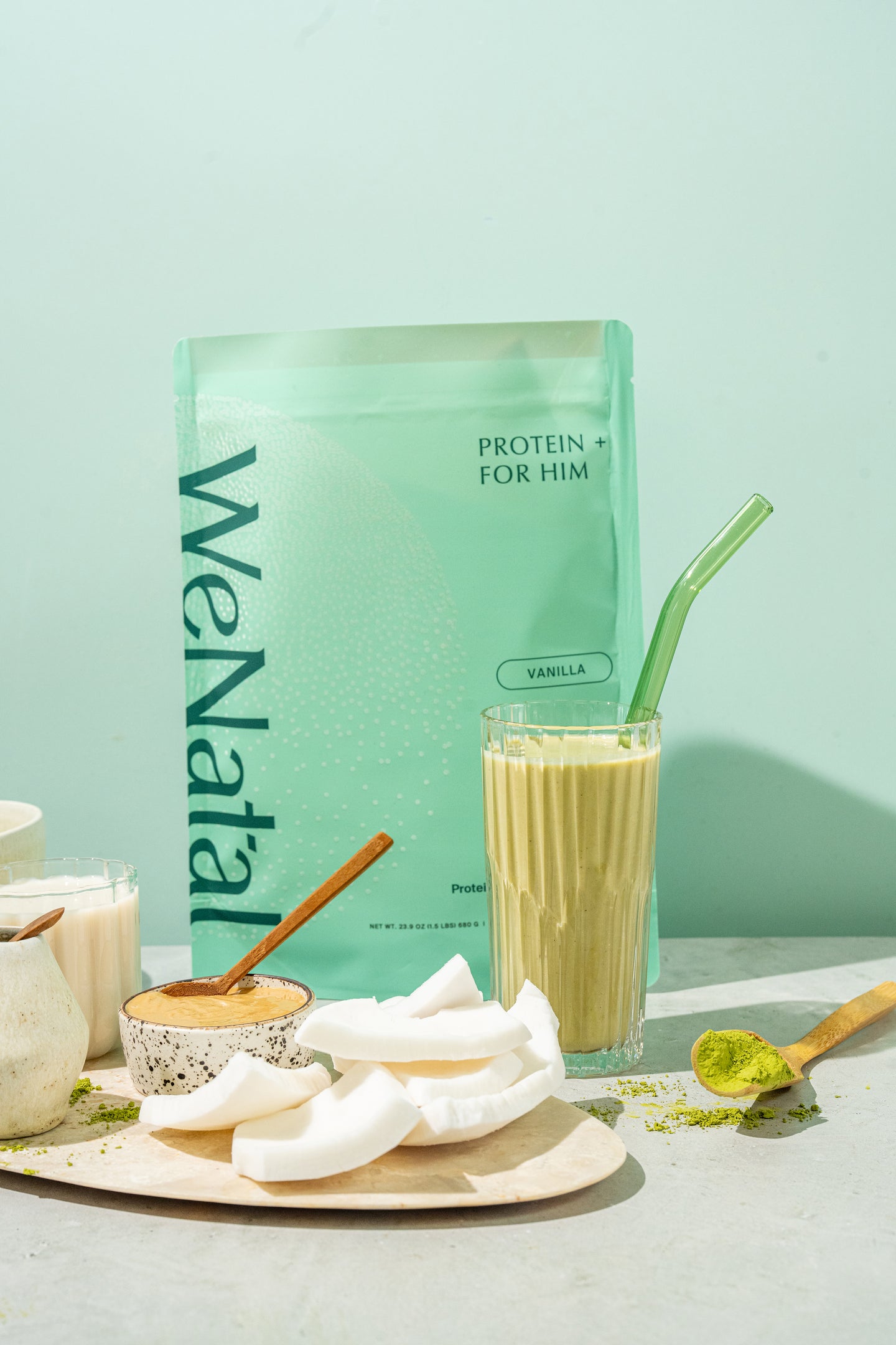 WeNatal Protein + For Him pouch with a vanilla flavor, surrounded by a green smoothie, coconut slices, nut butter, and matcha powder, conveying a health-focused vibe.