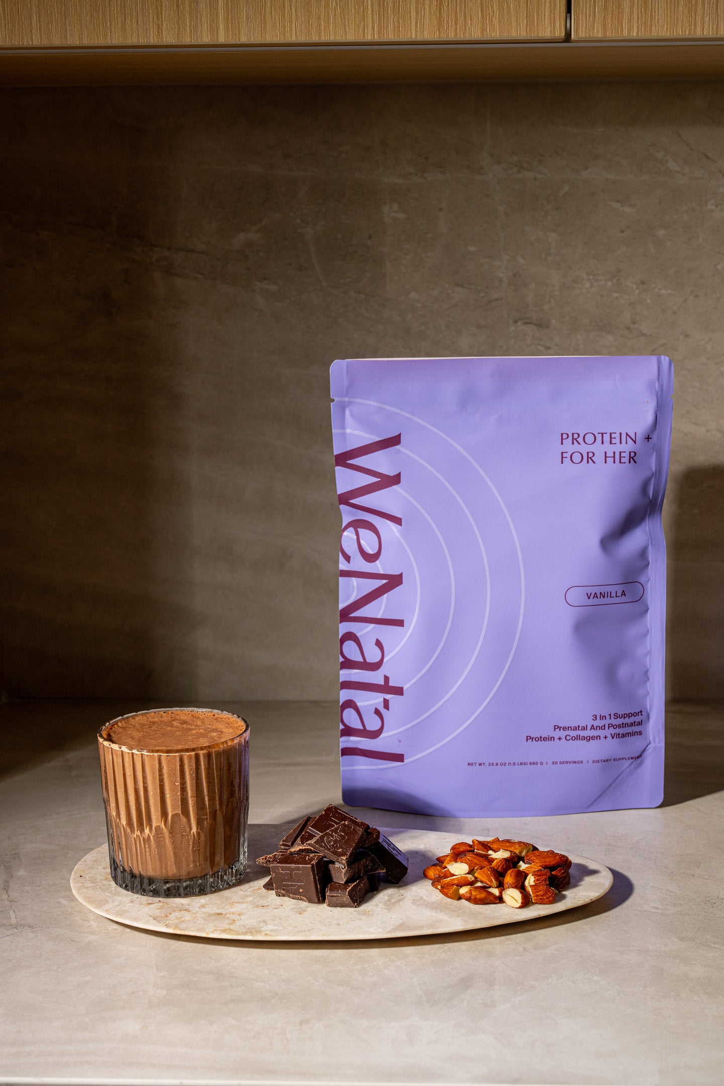 WeNatal Protein+For Her pouch with a chocolate shake in a fluted glass, surrounded by dark chocolate pieces and almonds on a marble board.