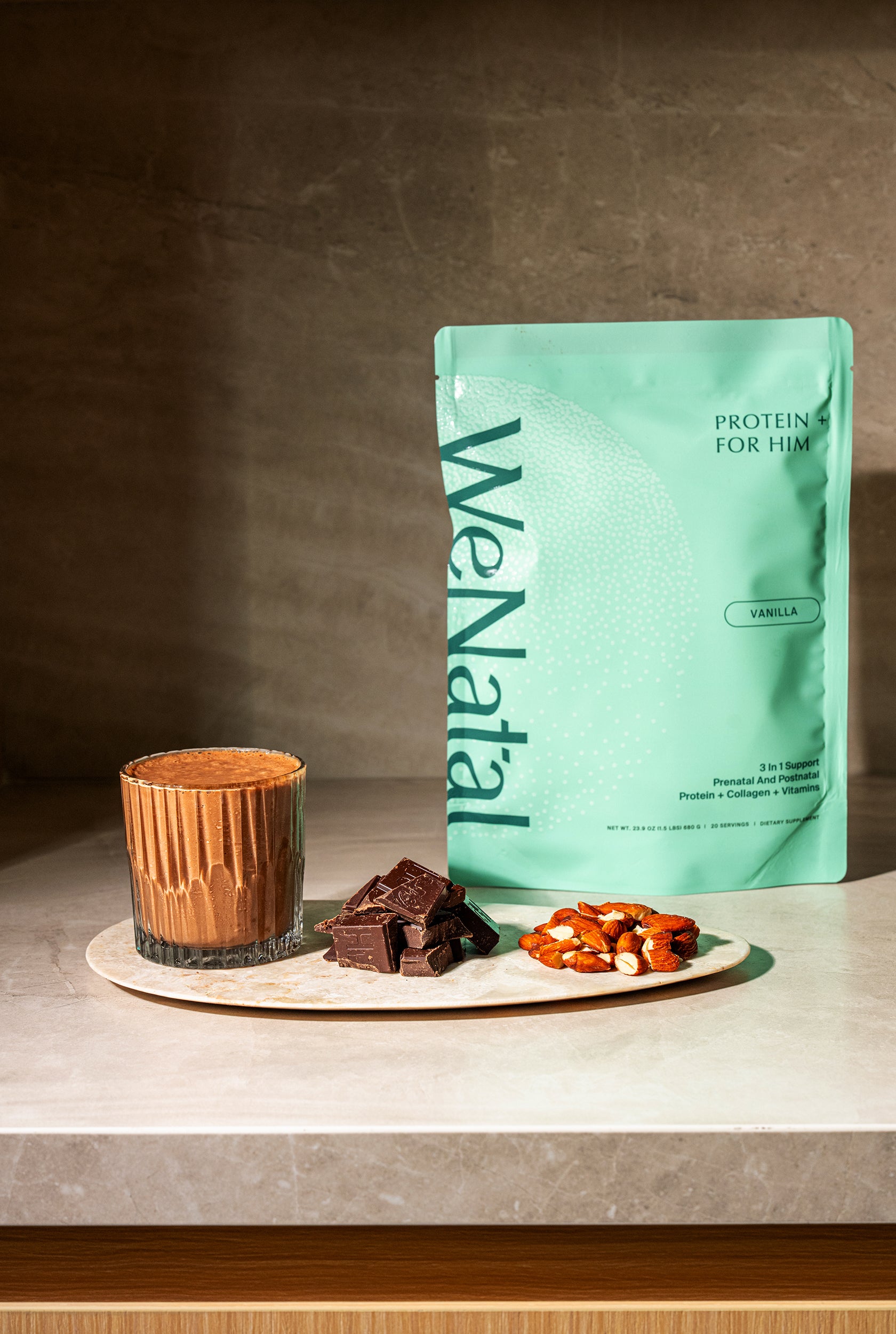 WeNatal Protein + For Him packet with a chocolate shake, dark chocolate chunks, and almonds on a marble board, highlighting the protein supplement's flavor profile