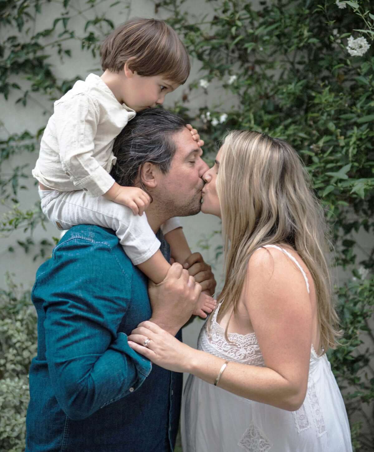 photo of a couple kissing, the girl is pregnant while the guy has their son sitting on his shoulders. the sone kissing the dad's head
