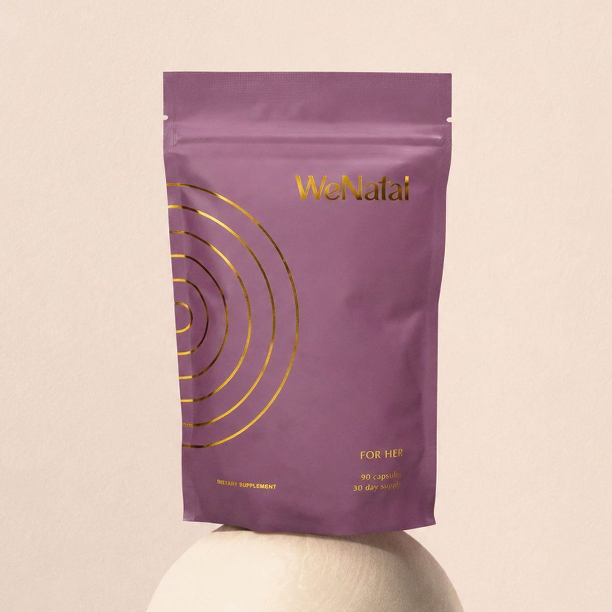 A purple WeNatal For Her pouch with 90 capsules for a 30-day supply, standing on a pedestal against a soft pink background.